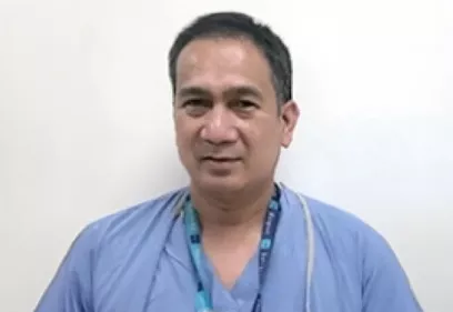 A head-and-shoulders portrait of a person wearing light blue medical scrubs and an ID lanyard. He is facing the camera with a slight smile against a plain light-colored background, creating a simple clinical or hospital setting.