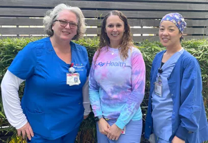Three nurses stand outdoors in front of greenery, smiling at the camera. The nurse on the left wears blue scrubs with an ID badge, the person in the center wears a colorful UCSF Health tie-dye sweatshirt and light-colored pants, and the nurse on the right wears blue scrubs with a patterned surgical cap. The group poses together in recognition of Certified Nurses Day.