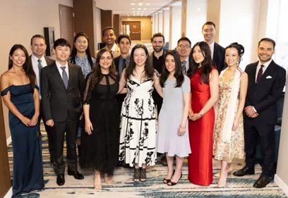 Group portrait of graduates and faculty dressed in formal attire, smiling together in a hotel hallway during a commencement celebration.