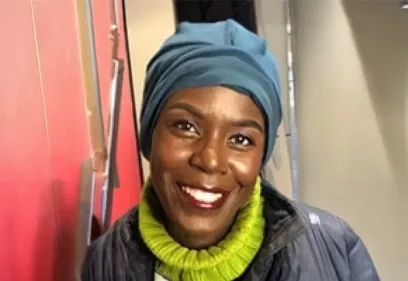 A head-and-shoulders portrait of a smiling person indoors. She is wearing a blue headwrap, a bright green turtleneck sweater, and a dark quilted jacket. She is looking directly at the camera, with a softly lit interior and red accent wall in the background.