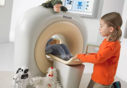 A young child kneels beside a child-friendly CT scanner, gently sliding a plush toy onto the scanner bed for a pretend imaging exam. The scanner is decorated with a stuffed crocodile on top, while a plush elephant rests on the scanner bed. A toy robot and stuffed chicken sit on the floor nearby. The bright, welcoming room includes colorful educational posters, creating a playful environment designed to help children feel more comfortable with medical imaging.