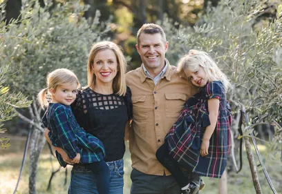 Family of four smiling and posing together outdoors among olive trees, with the parents standing between their two young daughters.