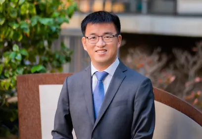 Professional portrait of a smiling man wearing glasses, a gray suit, white shirt, and blue tie, standing outdoors in front of greenery and a blurred building.