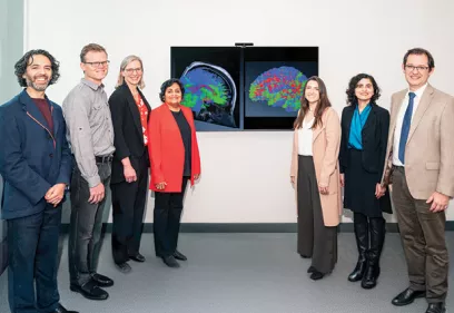 Seven people stand together in a clinical or research setting beside two wall-mounted monitors displaying colorful brain imaging scans.