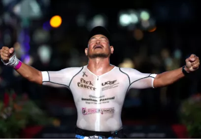 Athlete crossing a finish line at night with arms raised in celebration, wearing a white racing suit displaying cancer awareness and UCSF-related logos.
