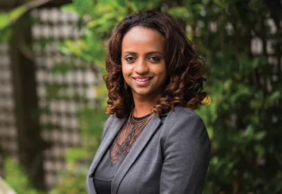Woman in professional attire smiling outdoors with greenery in the background.