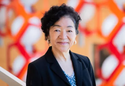 A woman in a dark blazer smiles at the camera, standing in front of a colorful red and orange geometric background.