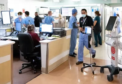 Healthcare workers in scrubs and masks work around a busy hospital nursing station with computer workstations and medical equipment.