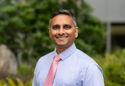 Portrait of a smiling man wearing a light blue dress shirt and coral tie, standing outdoors with greenery in the background.