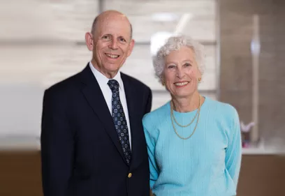 An older man in a dark suit and tie stands beside an older woman wearing a light blue top and gold necklaces. Both are smiling at the camera.
