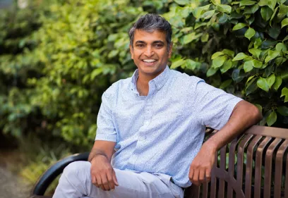 Man smiling while seated on a bench outdoors surrounded by greenery.