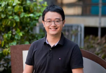 Man wearing glasses and a black polo shirt smiling outdoors with greenery in the background.