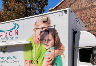 Mobile breast care and mammography van parked beside a brick building, featuring a large photograph of an older woman embracing a young girl on its side.