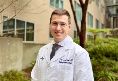 A man wearing glasses, a white UCSF medical coat, dress shirt, and tie stands outdoors on a landscaped medical campus, smiling at the camera.