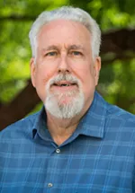 Professional headshot of a man with white hair and a neatly trimmed white beard, wearing a blue collared shirt, standing outdoors with greenery softly blurred in the background.