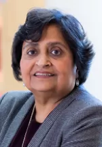 Professional headshot of a woman with short dark hair wearing a gray blazer, smiling gently at the camera against a softly blurred indoor background.