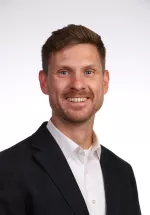 Professional headshot of Adrian Marth, a man with short brown hair and a trimmed beard wearing a dark blazer and white shirt against a light gray background.