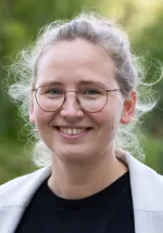 Portrait of Katharina Ziegeler smiling outdoors, wearing round glasses and a light gray jacket with a blurred green background.