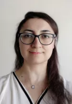 Headshot of Zehra Akkaya, a woman with dark hair wearing black glasses and a white blouse against a light background.