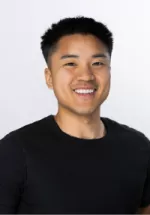 Smiling man with short black hair wearing a black t-shirt against a light gray background, facing the camera in a professional headshot.