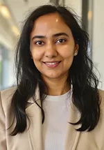 Headshot of a woman with long dark hair, smiling, wearing a light-colored blazer and top, against a softly blurred indoor background.