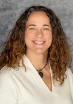 Portrait of a smiling woman with shoulder-length curly brown hair, wearing a light beige blouse and a black cord necklace with a dark pendant, set against a soft gray studio background.