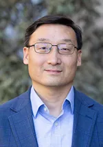 Portrait of a man wearing glasses and a blue blazer over a light blue shirt, standing outdoors with a softly blurred natural background.