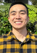 Headshot of a smiling man with short dark hair, standing outdoors in bright sunlight. He is wearing a yellow and black plaid button-up shirt. Green foliage fills the background, creating a natural setting with soft shadows and vibrant colors.