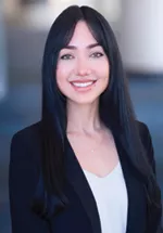 Professional headshot of a smiling person with long dark hair wearing a black blazer and white blouse against a softly blurred indoor background.