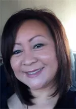 Head-and-shoulders portrait of a smiling woman with shoulder-length dark hair, wearing light makeup and looking toward the camera. The photo is taken indoors with a softly blurred background.