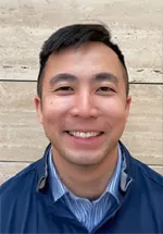 Professional headshot of a smiling person with short dark hair wearing a blue zip-up jacket over a blue striped collared shirt. The subject is facing the camera and standing in front of a light beige stone wall. The portrait is well-lit and framed from the shoulders up, conveying a friendly and approachable appearance.