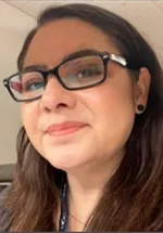 Head-and-shoulders portrait of a woman wearing rectangular glasses and small earrings, smiling softly toward the camera. She has shoulder-length dark hair and is photographed indoors with a blurred background.
