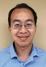 Head-and-shoulders portrait of a man wearing glasses and a light blue collared shirt, smiling gently at the camera. The photo is set against a neutral beige background.