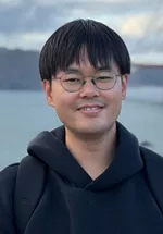 Portrait of a man wearing glasses and a dark hoodie, smiling slightly while standing outdoors. A body of water and cloudy sky are visible in the softly blurred background.