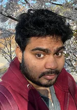 Headshot of a man with dark curly hair and short facial hair, looking toward the camera with a slight smile. He is wearing a maroon hoodie over a gray shirt and a necklace. The photo was taken outdoors, with flowering tree branches and a softly blurred background behind him.