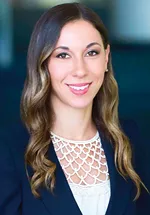 Professional headshot of a smiling person with long wavy brown hair wearing a navy blazer and cream blouse against a softly blurred blue background.