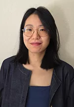 Portrait of a person with shoulder length dark hair and round glasses, wearing a navy blue jacket over a blue top. They are smiling slightly while facing the camera against a plain light colored wall.