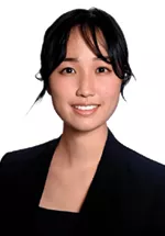 Professional headshot of a smiling person with dark hair tied back, wearing a black blazer and black top against a white background.