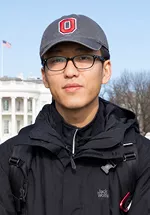Portrait of a person wearing black framed glasses, a gray baseball cap with a red and white "O" logo, and a black jacket with backpack straps visible. They are facing the camera with a neutral expression outdoors, with the White House and an American flag visible in the background.