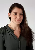 Alt text:  Portrait of a person with long dark brown hair wearing a dark green blouse and layered necklaces. They are facing the camera with a slight smile against a plain light gray background.