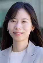 Head-and-shoulders portrait of a woman with long dark hair, smiling at the camera. She is wearing a light-colored blazer over a white top, with a softly blurred outdoor background.