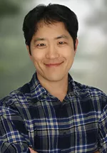 Professional headshot of a smiling person with short black hair, wearing a blue plaid button-down shirt. The background is softly blurred in shades of gray and green, keeping the focus on the subject.