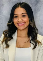 Professional headshot of a smiling person with long, dark wavy hair featuring light highlights, wearing a cream blazer over a white top, against a blue-gray studio background.