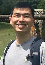 Outdoor head-and-shoulders photo of a person smiling at the camera, wearing a gray T-shirt and a black backpack. A stone railing and a pond with greenery are visible in the background.