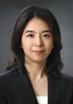 Professional headshot of a person with shoulder-length dark hair, wearing a black blazer over a light-colored top and a delicate necklace. The subject is smiling gently at the camera against a dark gray gradient background.