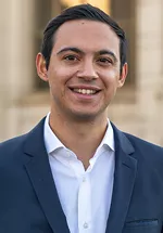 Head-and-shoulders portrait of a person smiling at the camera, wearing a navy blazer over a white collared shirt. The background is softly blurred, showing light-colored stone architecture.