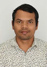 Professional headshot of a person with short dark hair, wearing a white button-down shirt with a blue floral pattern. The subject is facing the camera with a neutral expression against a plain light gray background.