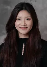 Professional headshot of a person with long, dark brown hair, wearing a black top with a decorative neckline, smiling gently at the camera against a gray studio background.
