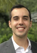 Headshot of Mateus wearing a gray blazer and white shirt, smiling outdoors with greenery in the background.