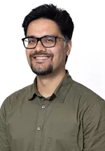 Headshot of a smiling person with short dark hair, glasses, and a goatee, wearing an olive-green button-up shirt against a light gray background.